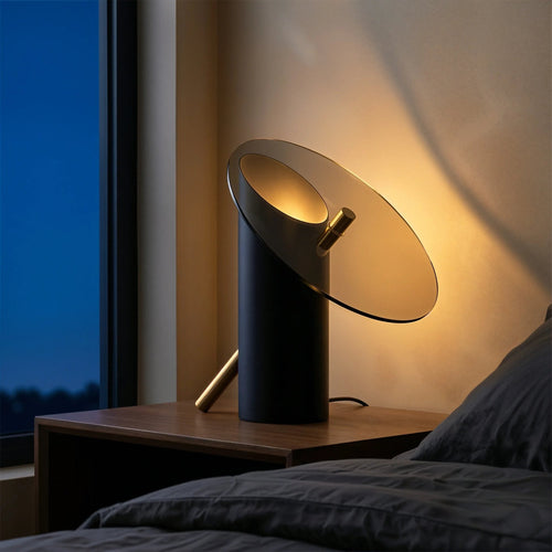 Obsidian bedside table lamp by Luminor Arc featuring a sleek architectural dark cylinder and an angled smoked glass disc, casting a warm amber glow against a textured wall during a deep blue hour.