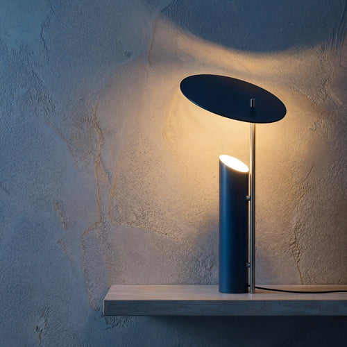 Velum table lamp by Luminor Arc featuring a minimalist architectural cylinder and an adjustable floating reflector, casting a warm amber glow against a textured plaster wall in a deep blue hour setting.