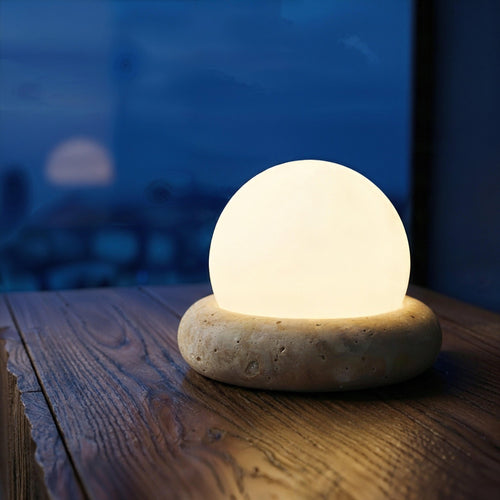 Nomad Sphere cordless lamp featuring a textured stone-like base and frosted globe, casting a warm amber glow on a wooden surface against a dark blue twilight window.