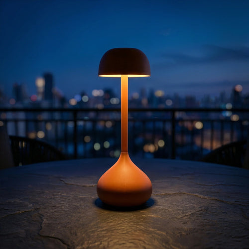 The Radix cordless lamp in matte leather orange aluminum, casting a warm amber glow on a textured stone table against a deep blue hour skyline.